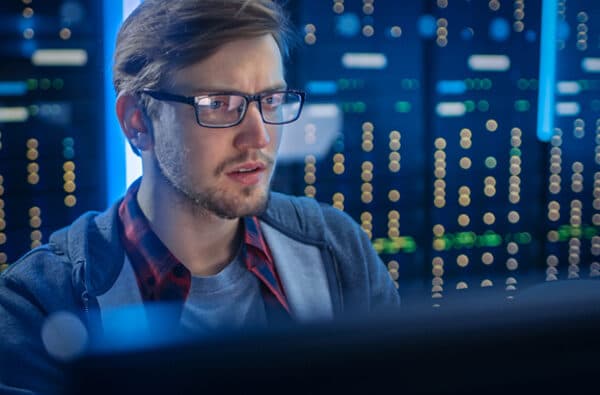 Portrait of a Smart Focused Young Man Wearing Glasses Ð¡oncentrated on a Desktop Computer. In the Background Technical Department Office with Functional Data Server Racks.